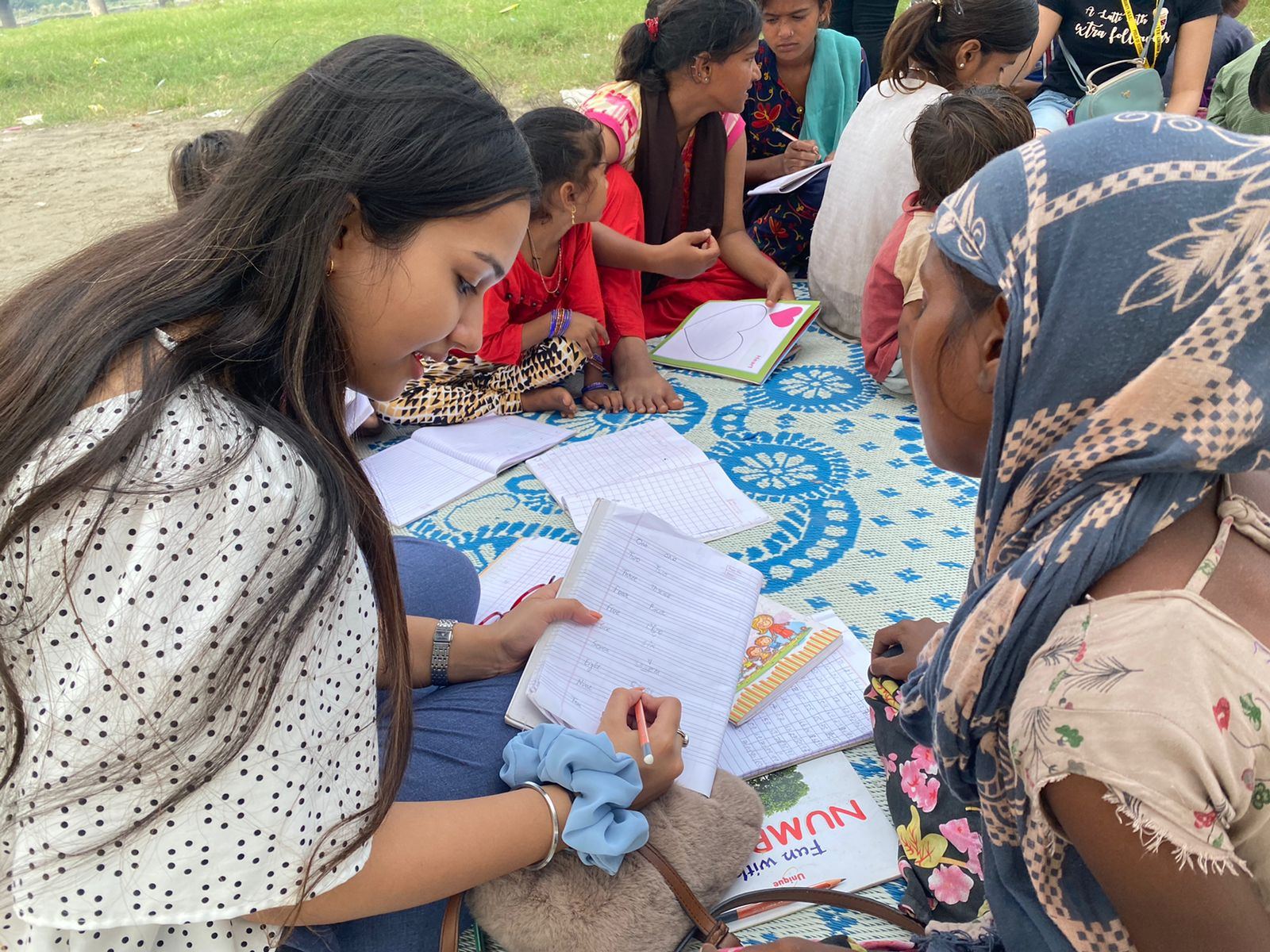 An open-air class on floor mats — a volunteer guides children through their notebooks